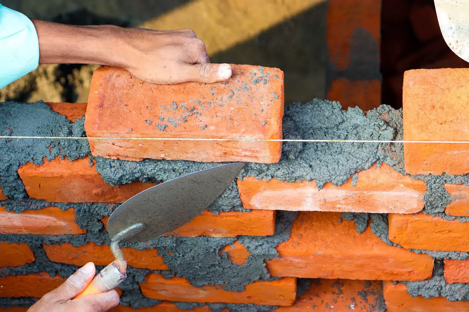 Bàn tay thợ xây đặt viên gạch lên lớp vữa, dây căng ngang giữ thẳng hàng tường