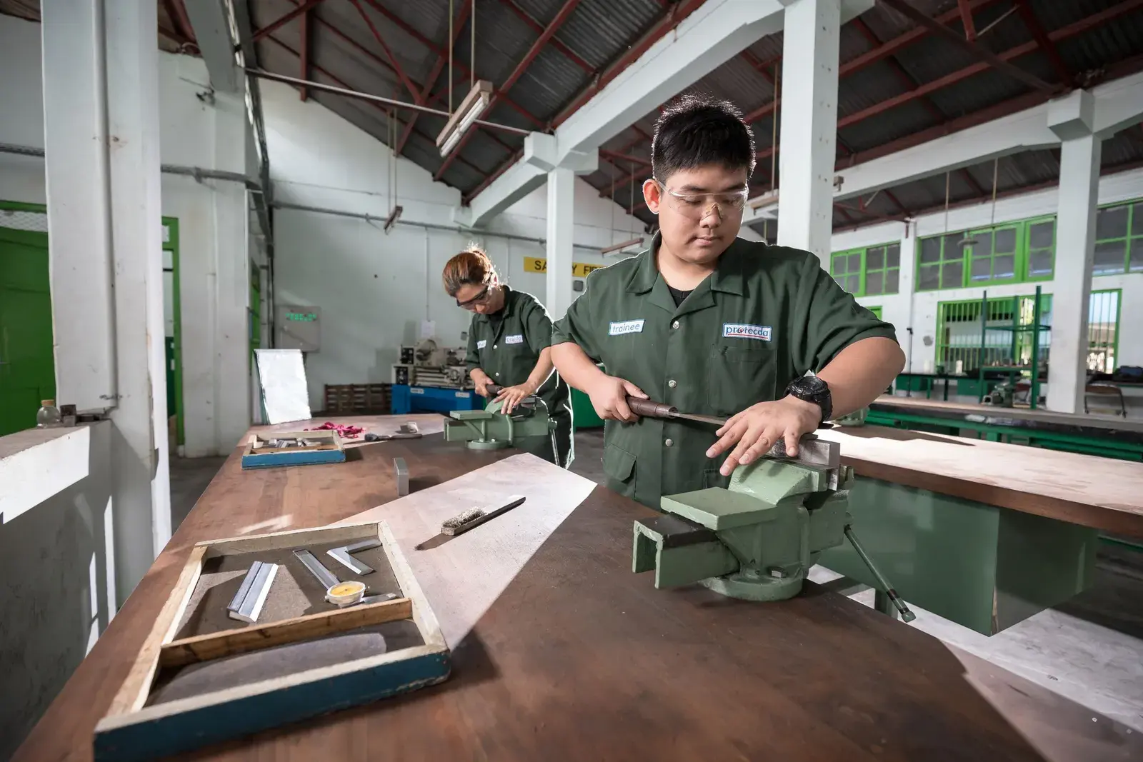 Học viên thao tác trên bàn nguội trong xưởng thực hành, giảng viên quan sát phía sau