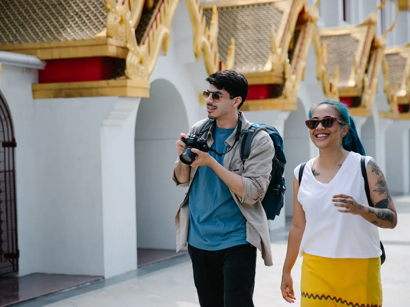 Hai du khách đeo ba lô, cầm máy ảnh, đi dọc hành lang một ngôi chùa mái vàng dưới nắng